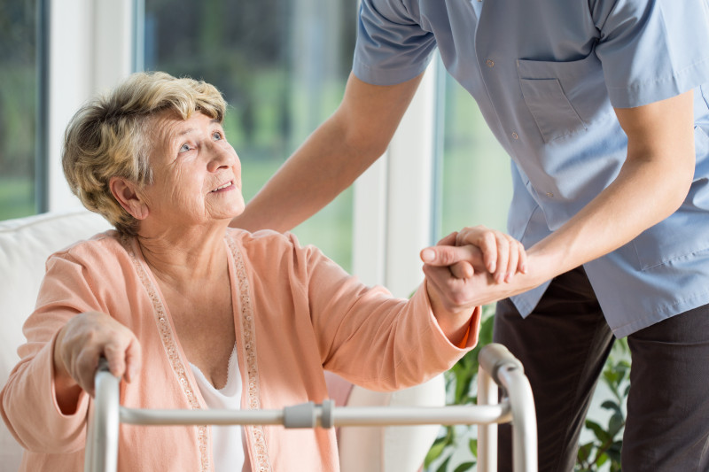Resident sitting with a walker in front of her holding hands and looking up at a nurse.
