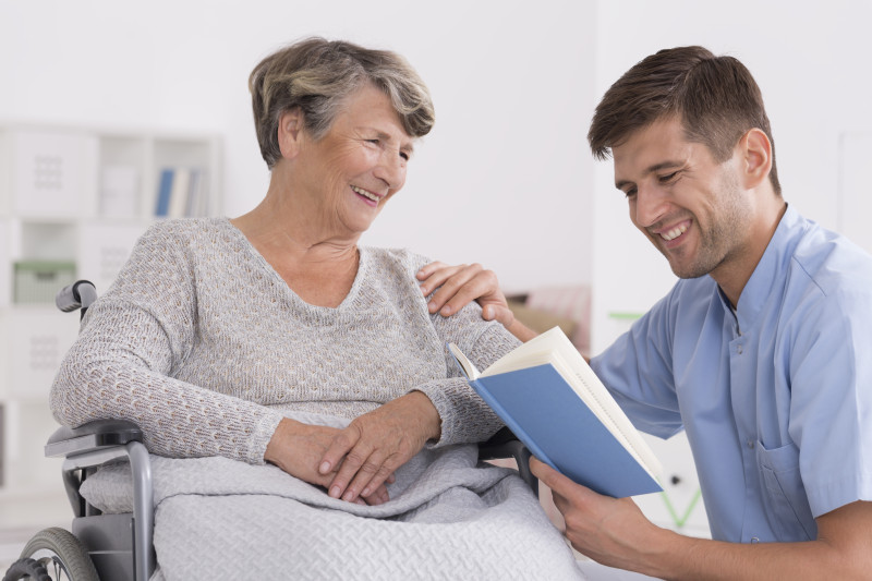 Resident sitting in a wheelchair while a nurse kneels next to her reading a book.