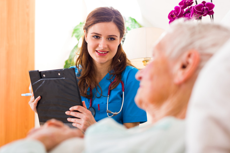 Resident laying in a hospital bed while a nurse with a clipboard talks to him.
