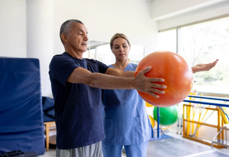 Patient holding a ball while a physical therapist helps him.